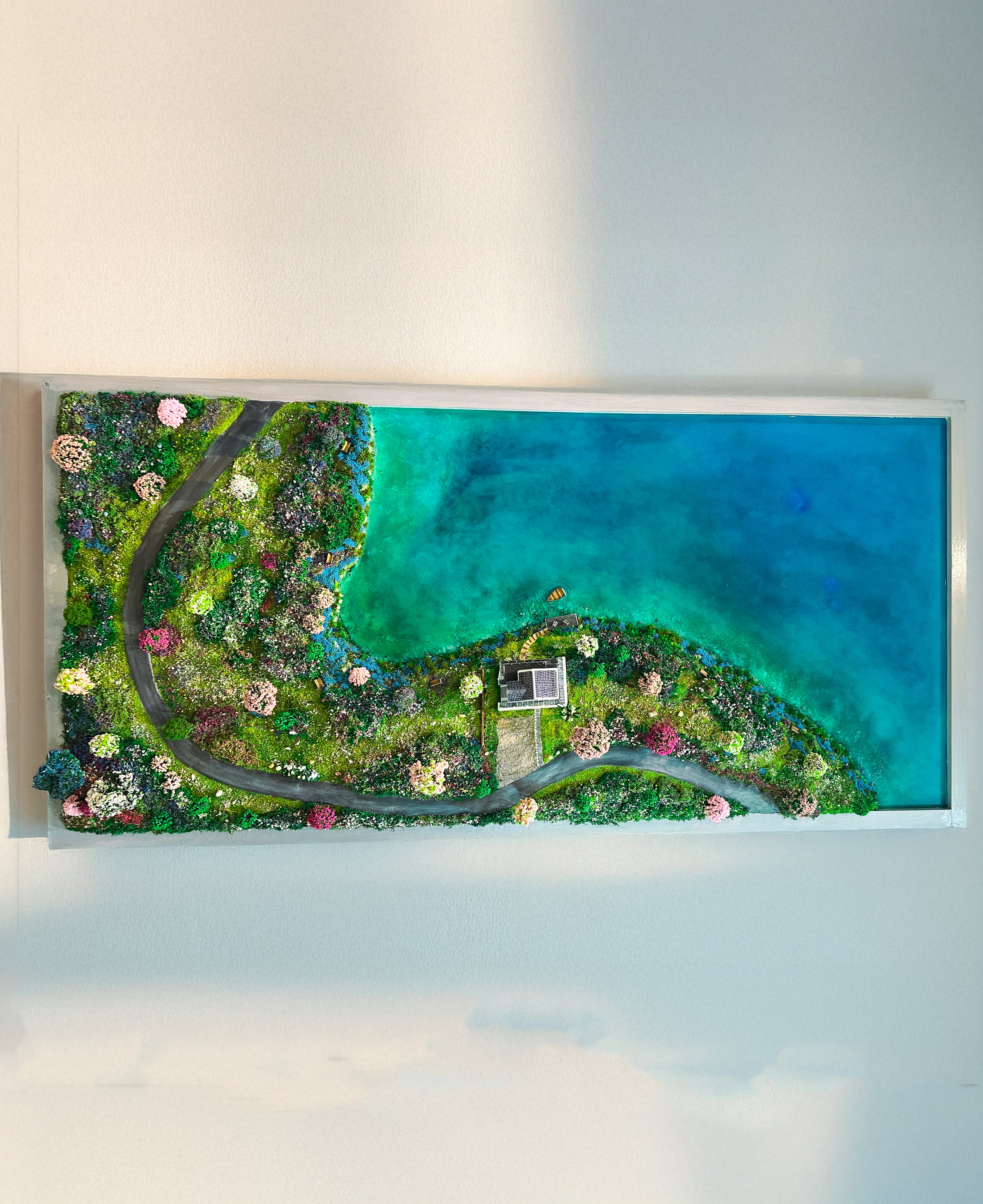 A framed landscape art print depicting a vibrant green coastline with a winding road, colorful flowers, and a bright turquoise ocean, mounted on a white wall.