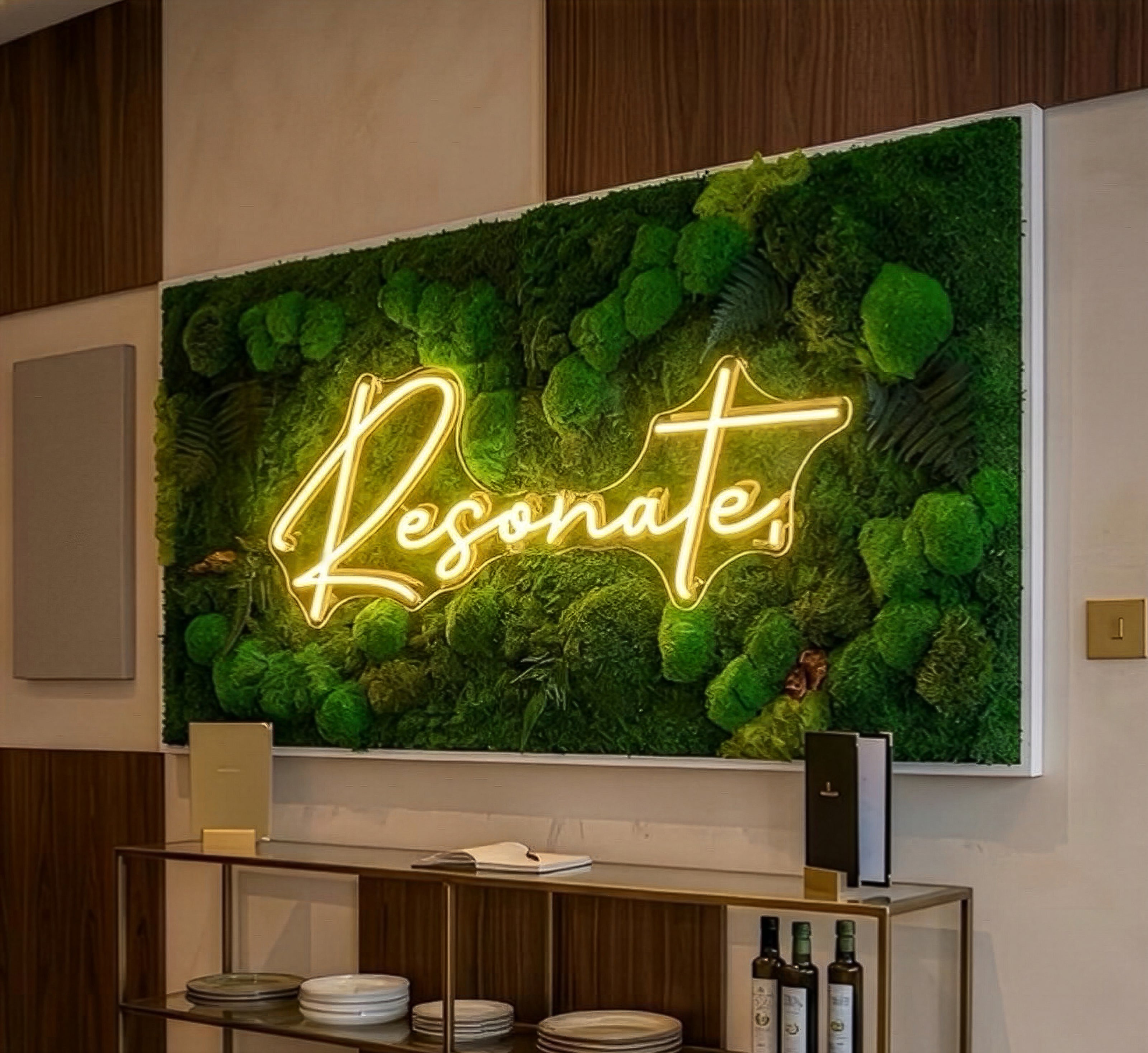 A large framed wall art piece featuring a green moss and fern background with a glowing yellow neon-style script reading “Resonate,” displayed in an interior setting above a shelf.