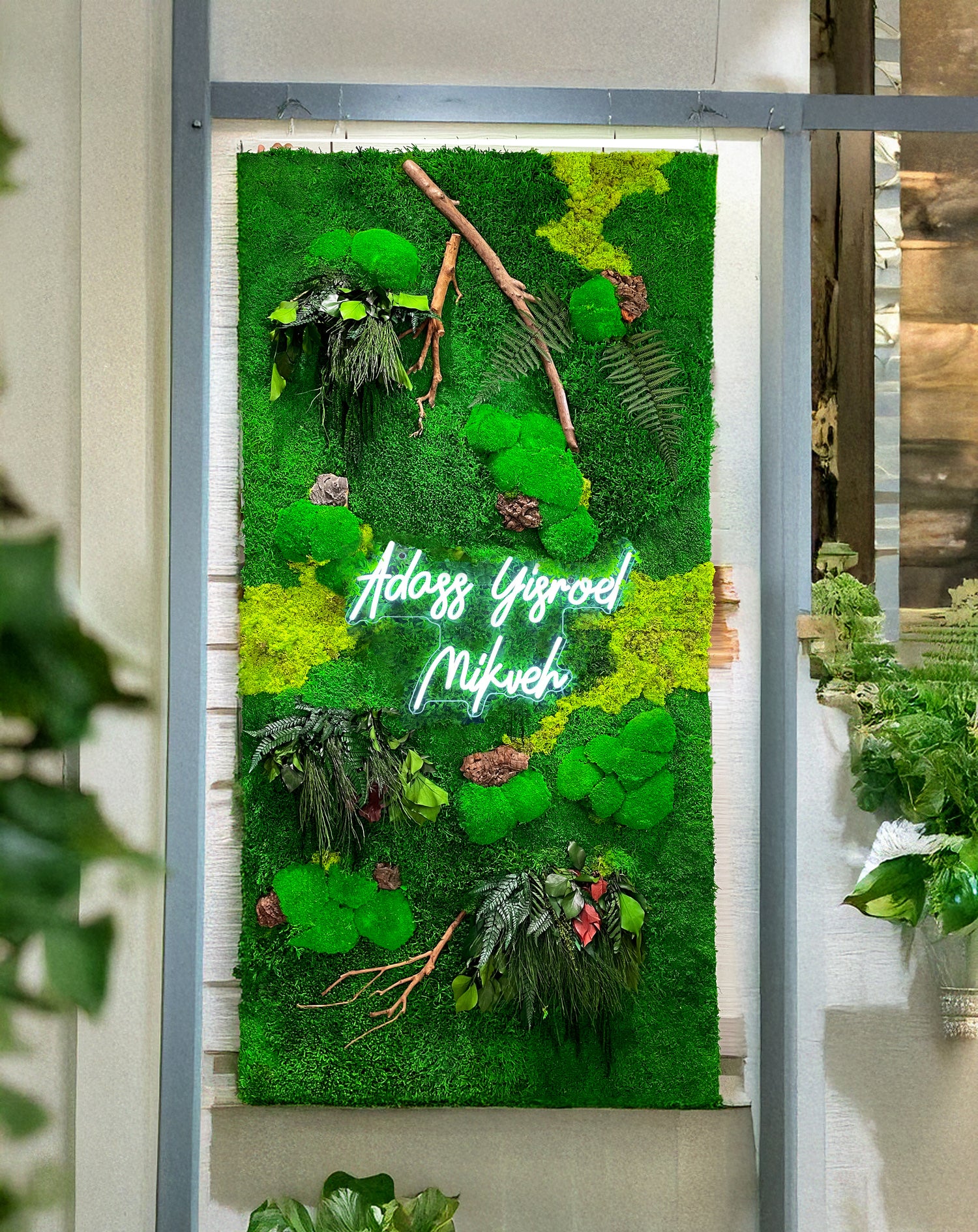 A vertical green wall hanging made of textured artificial grass, faux ferns, pinecones, twigs, and small floral accents, with the names 'Adams Yigroed' and 'Mikveh' in glowing white script across the center. The hanging is displayed indoors against a light wall within a gray frame.